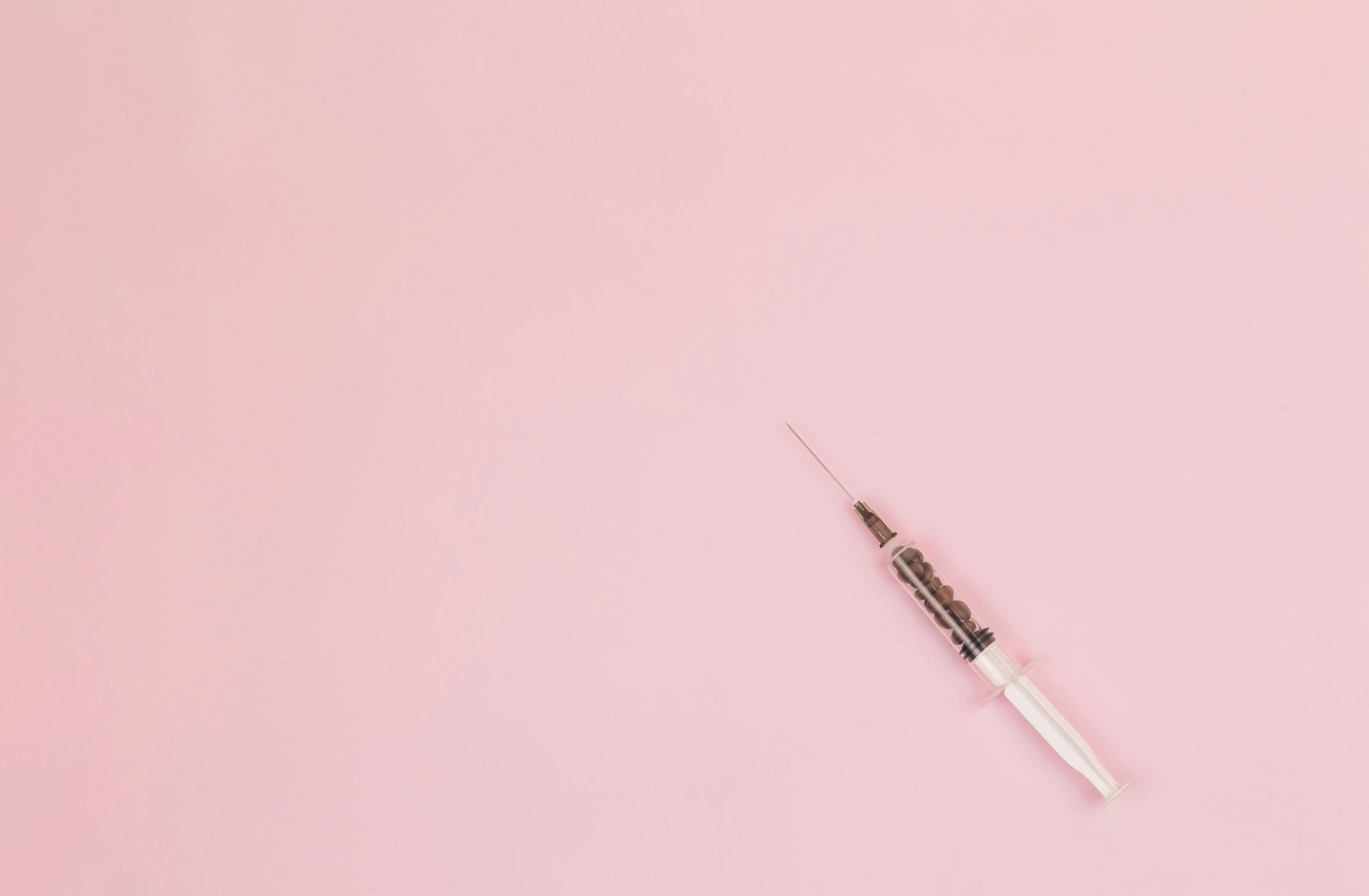 Medical syringe filled with dark coffee beans on a minimalist light pink background.